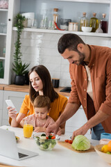 Man cooking salad near wife using smartphone and toddler son at home