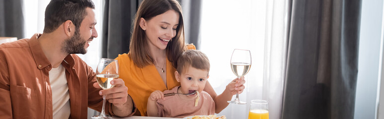 Smiling parents holding wine while son eating pasta at home, banner