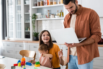 Man using laptop near smiling wife and son with building blocks at home