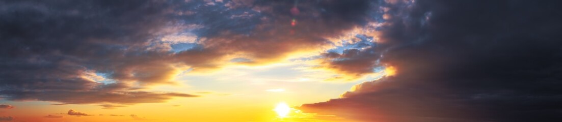 Panorama of dramatic colorful sunset with dark and bright clouds. The sun's rays are breaking through the cloudy sky. Dark clouds against a bright saturated sky.