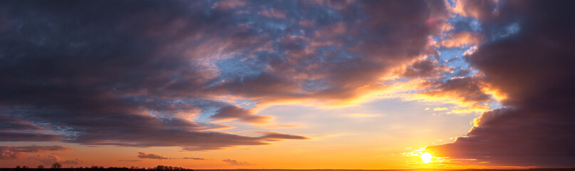 Panorama of dramatic colorful sunset with dark and bright clouds. The sun's rays are breaking through the cloudy sky. Dark clouds against a bright saturated sky.