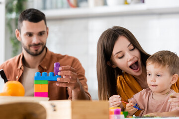 Woman holding paintbrush near son and husband with building blocks at home