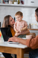 Happy mom hugging kid while husband using laptop at home