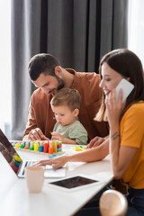 Obraz premium Smiling man sitting near son playing educational game and wife working on laptop and talking on cellphone at home