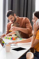 Smiling woman talking on smartphone and using laptop near blurred family at home