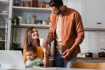 Smiling woman holding cup near husband, son and devices at home