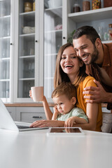 Happy man hugging wife with cup near son and gadgets at home