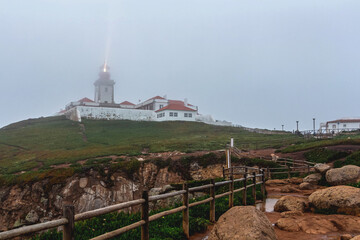 Fototapeta na wymiar A landscape with a luminous lighthouse in cloudy weather on Cape Roca in Portugal, a wooden fence, behind which there are greenery with flowers and brown soil with large stones and a puddle on the pat