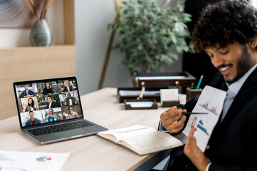 Positive successful Indian businessman, top manager or coach, uses laptop for online conference with employees, holds briefing, shows documents and charts, on screen are multiracial business people