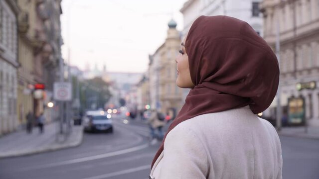 A young beautiful Muslim woman looks around with a smile in a street in an urban area - a road in the blurry background - closeup from behind