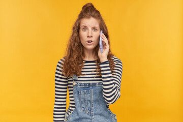 indoor studio shot of young ginger female with long red curly hair talking on the phone with confused, guilty facial expression, isolated over yellow background