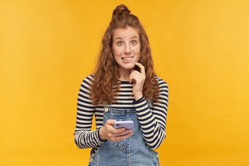 indoor studio shot of young ginger female with long red curly hair biting her lip, holding phone with ashamed guilty facial expression, isolated over yellow background - obrazy, fototapety, plakaty