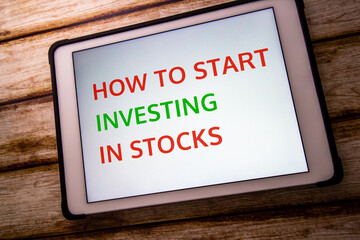 Conceptual keyword “How to Start Investing in Stocks” on tablet on faded shabby table....
