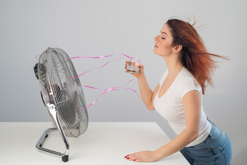 A red-haired Caucasian woman chills by the electric fan and drinks a cold drink. Climate control in...