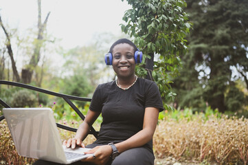 Smiling happy African black short-haired woman student afro hair with blue headphones studying online working on laptop computer at summer green park. Diversity. Remote work, distance education