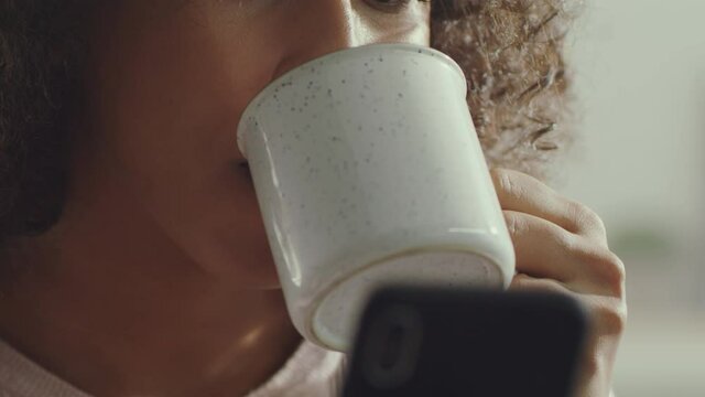 Close up portrait of unrecognizable african american woman drinking tea or coffee and web surfing online on cellphone