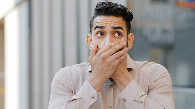 Confused afraid scared young guilty man shocked surprised guy male hispanic arab indian covers mouth with hands worried about telling secret feels shock bad news looking at camera, problem concept