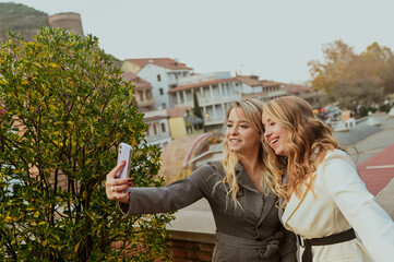 Close-up portrait of two female friends in strict suits laughing taking selfie on the terrace outside at summer street cafe on background buildings of Old Tbilisi city, Georgia. High quality photo