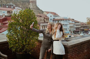 Close-up portrait of two female friends in strict suits laughing taking selfie on the terrace outside at summer street cafe on background buildings of Old Tbilisi city, Georgia. High quality photo