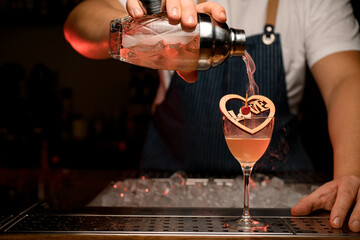 hand of bartender pours a steaming alcoholic cocktail from glassy shaker into wineglass with wooden decoration