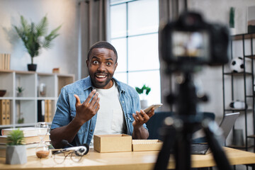 Happy african american man testing new modern smartphone while recording video on camera at home. Male influencer doing live stream. People and social networks concept.