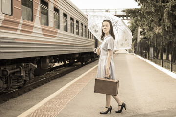 An elegant lady in a dress with an umbrella and a vintage suitcase is waiting for her train on the...