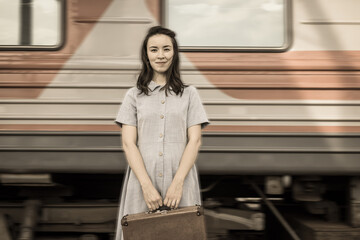 A young woman in a dress and carrying a suitcase is standing on a platform with a blurry fast-moving train in the background. Retro-style processing. The concept of traveling by train.