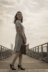 A young brunette woman in a dress with a vintage suitcase in her hands stands against a clear blue sky. Retro style, vintage. Peaceful sky.