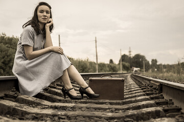 A young woman in a dress with a vintage suitcase is sitting on the rails on a railway track. The...