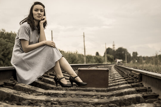 A young woman in a dress with a vintage suitcase is sitting on the rails on a railway track. The concept of travel.