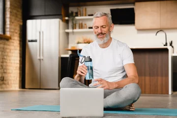 Wandcirkels plexiglas Closeup portrait of middle-aged handsome mature man in sporty outfit sitting in yoga lotus position on fitness mat watching video training tutorial on laptop after workout at home. © InsideCreativeHouse
