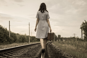 A rear view of a young woman in a dress and with a suitcase walking away along the rails of a...