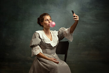 Retro portrait of young romantic girl in gray dress of medieval style using phone isolated on dark vintage background.