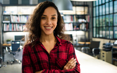 Beautiful cheerful smart hipster Caucasian creative woman wearing casual shirt, smiling with happiness, success, crossing arms, looking at camera, standing in modern indoor office. Business Concept.