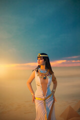 Egypt Style Rich Luxury Woman. Sexy beautiful girl goddess Queen Cleopatra stands, yellow sand desert pyramids. Art ancient pharaoh costume white dress gold accessories. Black hair wig Egyptian makeup