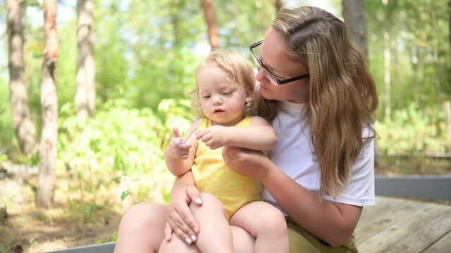 Little cute baby toddler girl blonde with curls on mother's arms. Mother and daughter playing spend time together outdoors at countryside front yard summertime. Healthy happy family childhood concept