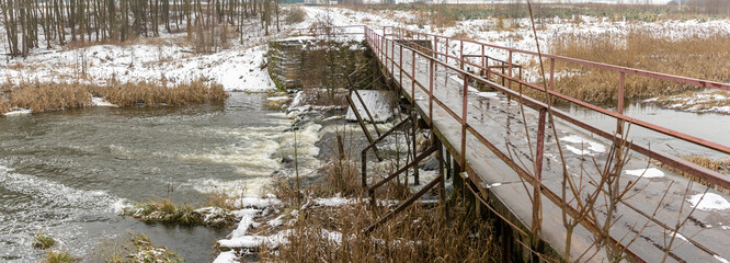 The narrow old wooden bridge over winter river. Wooden pedestrian bridge over river in countryside. Winter landscape.