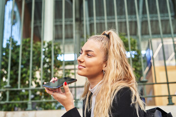Young female recording audio message on smartphone near modern building