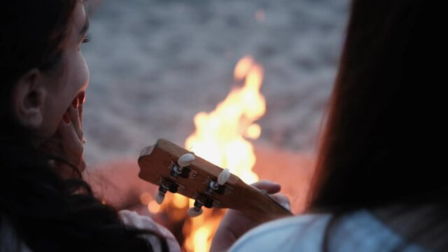 Young brunette girl plays by the fire on the slope. Girls are sitting on the beach by the fire. Summer holidays. Evening on the beach by the sea. Party of a young company. Friendly guys are resting on