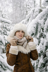 Fototapeta na wymiar Vertical winter portrait of a smiling young european girl in a sheepskin coat, mittens, a white hat with earflaps and boots, who stands in snowy pine trees