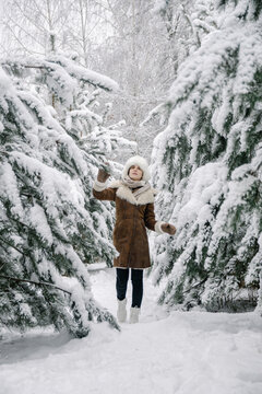 Vertical photo of a young European girl in a sheepskin coat, mittens, a white hat with earflaps and boots, walking among a pine forest in the snow in winter