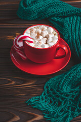 Red cup of cocoa with marshmallows and candy cane. Hot chocolate and warm knitted scarf on a dark wooden table. Winter season, card, minimal design. Christmastide, Saint Nicholas Day. New Year morning