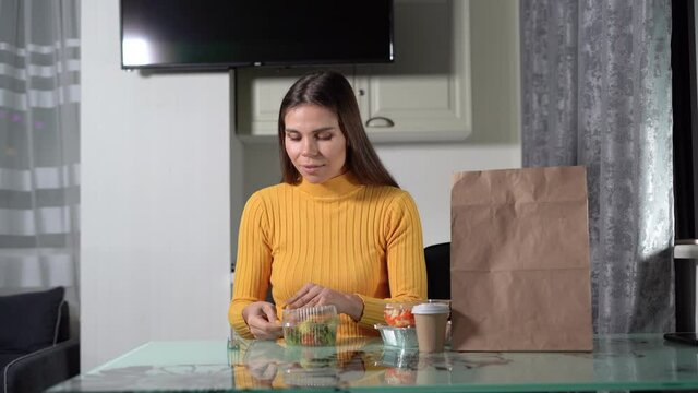 home delivery food concept. A young woman sits at the table in the evening, opens a plastic tray with food and begins to eat a vegetable salad.