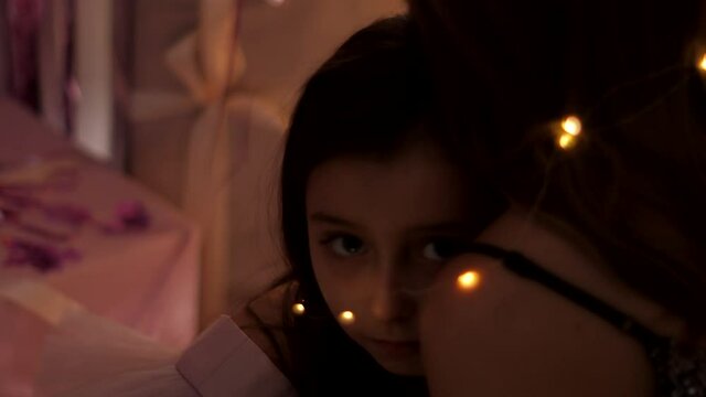 girl lies on her mother's shoulder tied with a garland and looks into the camera at night on Christmas day