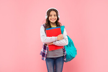 glad kid listening music in earphones with school bag and workbook on pink background, ebook