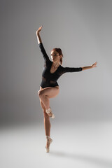 full length of graceful ballerina in black bodysuit dancing with outstretched hands on dark grey