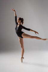 full length of elegant ballerina in black bodysuit dancing with raised hand on dark grey