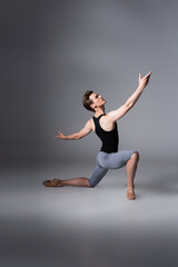 Fototapeta na wymiar young ballet dancer in black tank top performing ballet dance while standing on knee on dark grey