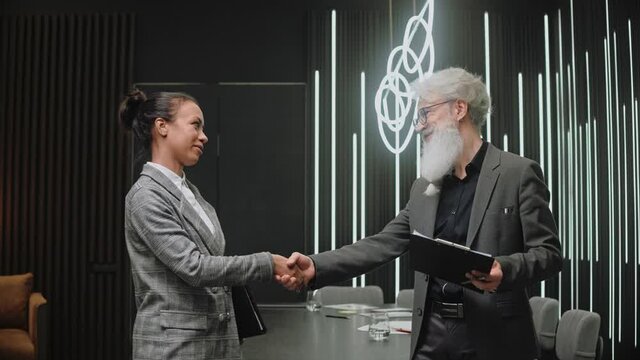Young girl talks with elderly businessman and shakes hands in office. People in suits discuss something in modern black office. High quality 4k footage.50 fps