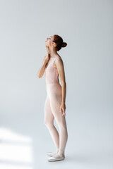 full length of elegant ballerina in ballet shoes touching neck on grey